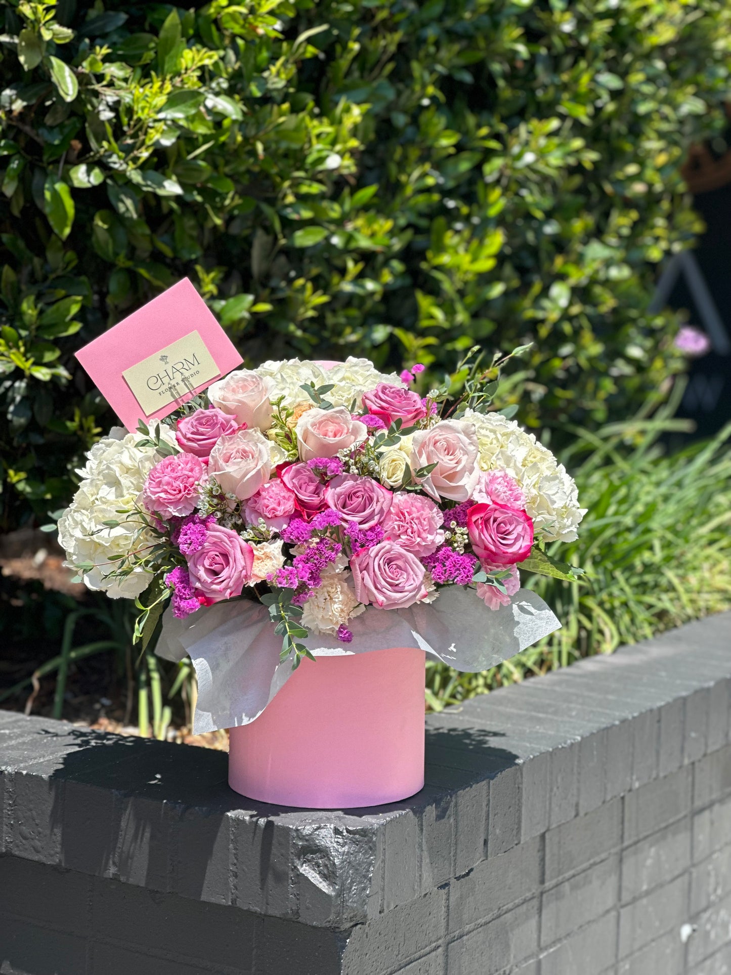 Soft pink hatbox filled with blush and hot pink roses, white spray roses, and seasonal accents. Flower selection and wrapping may vary slightly to ensure freshness.