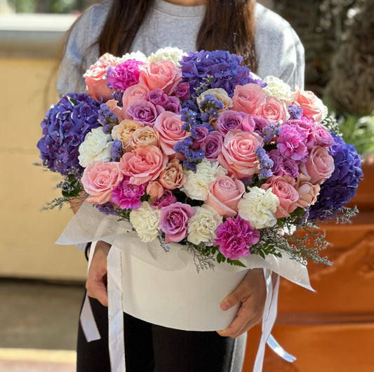 Luxurious mix of pink, lavender, and purple roses with blue hydrangeas, arranged in a hatbox. Colors and flower types may vary slightly to ensure freshness.